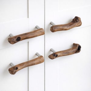 May include: Four wooden cabinet pulls with a natural, organic aesthetic. Each pull is crafted from a piece of wood, featuring a unique shape and texture, and is mounted on a brushed metal rod. The pulls are a warm brown color and are set against a white background.