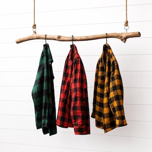 May include: Three plaid shirts in green and black, red and black, and yellow and black hang from a natural wood branch suspended by rope. The shirts are button-down and have long sleeves. The background is a white, horizontal-slat wall.
