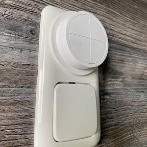 May include: White wall-mounted light switch with a round button on top. The button has four sections with small dots in each section.