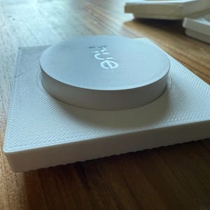 May include: A white, 3D-printed square base with a circular top. The top has the word "hue" printed on it. The base has a textured surface. Additional white objects are visible in the background.