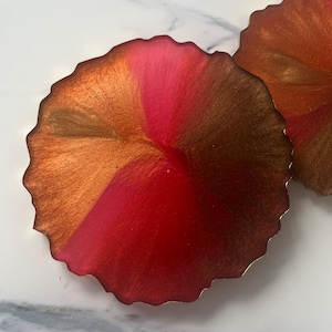 May include: Two round coasters with a distinctive design. They display a mix of red, pink, and gold tones, producing a vibrant look. The edges have a wavy, organic shape, enhancing their artistic appeal. These coasters would be a great addition to any home.