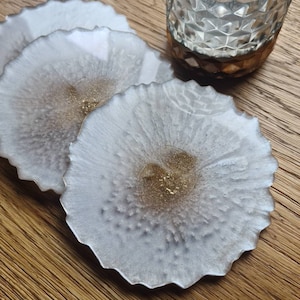 Könnte beinhalten: Drei weiße und goldfarbene Untersetzer aus Kunstharz mit einem strukturierten, organischen Design. Die Untersetzer haben einen gewellten Rand und eine goldene Mitte. Ein klares Glas mit braunem Boden steht daneben.