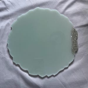 Op de afbeelding: Een ronde onderzetter van licht muntgroene hars met een onregelmatige, golvende rand. Eén kant van de onderzetter is voorzien van een textuurrijk zilveren glitteraccent dat de rand volgt en contrasteert met de gladde, matte afwerking van het oppervlak.