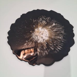 Peut inclure: Un dessous de verre en résine noire et dorée de forme organique. Le dessous de verre présente un bord noir brillant qui entoure un motif central d'accents blancs, dorés et argentés, créant un effet visuel saisissant. Le dessous de verre est sur une surface blanche.