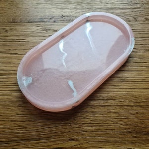 Könnte beinhalten: Ein rosa, ovales Tablett mit weißen Marmorierungen. Das Tablett ist aus Harz gefertigt und hat eine glatte, glänzende Oberfläche.