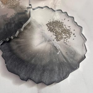 May include: Two circular coasters with irregular, jagged edges made of resin. The coasters feature a gradient design transitioning from clear to dark grey and black, with a cluster of small, metallic silver flakes embedded on the surface.