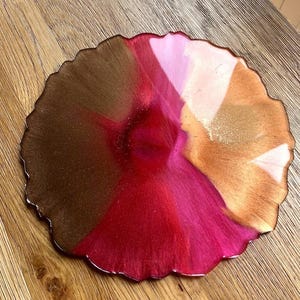May include: A round, decorative resin coaster with a unique, organic shape. The coaster features a blend of colors, including shades of brown, red, pink, and gold. The surface has a glossy finish, and the edges are slightly irregular.
