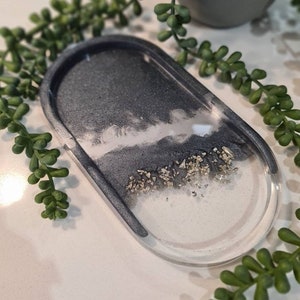 May include: A grey and white oval resin tray with a sparkly silver glitter design. The tray is ideal for holding jewellery, keys, or other small items.