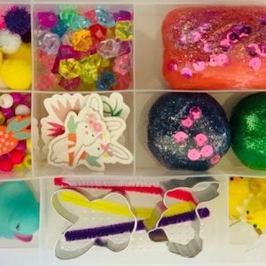 May include: A clear plastic storage box filled with various Easter-themed craft supplies. The box includes colorful pom-poms, beads, glitter glue, cookie cutters, and more.