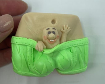 May include: A whimsical figurine featuring a smiling, cartoonish character peeking out of bright green underwear. The character has a light skin tone, rosy cheeks, and is waving. The underwear has a textured design.