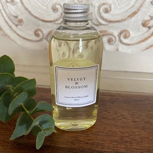 May include: A clear glass bottle with a silver lid filled with a light yellow liquid. The bottle has a white label with the text "Velvet & Blossom" and "Luxury Reed Diffuser Refill 100ml".