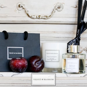 May include: A black gift bag with the text "Velvet & Blossom" printed on it, next to a white candle and a glass bottle of diffuser with black reeds. The candle and diffuser both have the text "Velvet & Blossom" and "Black Plum & Fig with Rosewood" printed on them. There are two dark red plums in front of the bag.