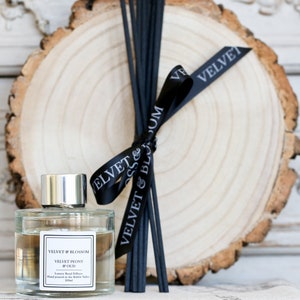 May include: A clear glass bottle of diffuser oil with a black label that reads "Velvet & Blossom Velvet Peony & Oud Luxury Reed Diffuser Hand poured in the Ribble Valley 100ml". The bottle is sitting on a wooden slice with black ribbon tied around it and black diffuser sticks.