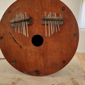 May include: A wooden kalimba, a type of thumb piano, with a circular soundboard. The instrument features two sets of metal tines and a central sound hole. The wood has a warm, reddish-brown hue.