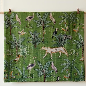 May include: Green textile with a jungle scene featuring palm trees, birds, a leopard, and a zebra. The design is printed on the fabric and has a quilted texture. The textile is hanging from a wooden rod with clips.
