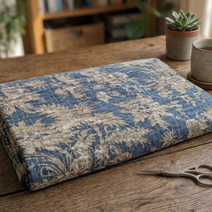 May include: A folded rectangular quilt featuring a blue and beige floral pattern with visible white stitching throughout. The fabric displays a dense botanical print of leaves and flowers, creating a textured appearance across the entire surface.