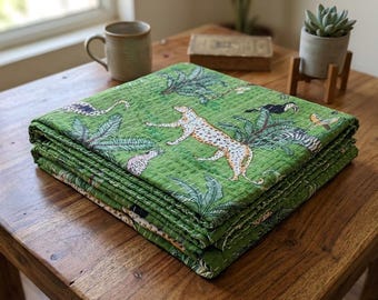 May include: A folded green quilt with a jungle-themed design featuring leopards, toucans, and tropical foliage. The quilt is stacked on a wooden table, showcasing its detailed pattern and vibrant colors.