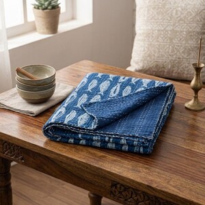 May include: A folded blue quilt featuring a white fish pattern and visible white stitching. Beside the quilt sits a stack of three small ceramic bowls on a light-colored cloth, and a brass incense holder stands on the wooden surface.