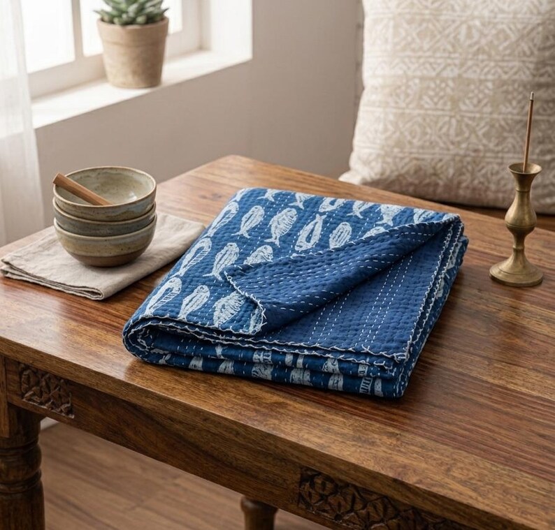 May include: A folded blue quilt featuring a white fish pattern and visible white stitching. Beside the quilt sits a stack of three small ceramic bowls on a light-colored cloth, and a brass incense holder stands on the wooden surface.