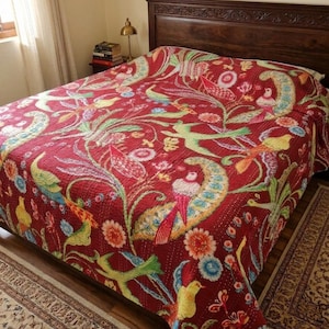 May include: A red quilt with a vibrant floral and bird pattern. The design features colorful birds, flowers, and foliage in shades of green, yellow, blue, and orange. The quilt is spread over a wooden bed frame.