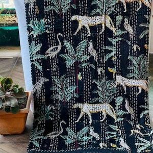 May include: A black kantha quilt featuring a repeating pattern of white leopards, zebras, and long-necked birds interspersed with green palm trees. The fabric is decorated with rows of white running stitches that create a textured, quilted appearance.