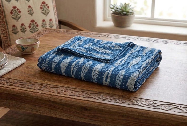 May include: A folded rectangular quilt featuring a deep blue background with a repeating white fish pattern. The fabric displays visible white running stitch details across the surface, creating a textured appearance throughout the textile piece.