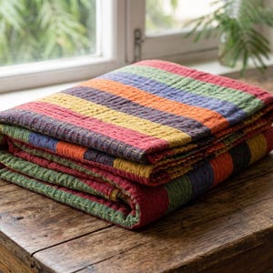 May include: A folded rectangular quilt featuring horizontal stripes in vibrant shades of red, yellow, orange, green, and blue. The fabric has a visible quilted texture with fine, light-coloured stitching running across the surface of the textile.