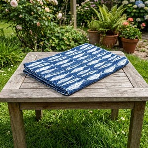 May include: A folded rectangular textile featuring a repeating pattern of white fish silhouettes on a dark blue background. The fabric has a visible quilted texture and is positioned flat on a square wooden table with a slatted surface design.