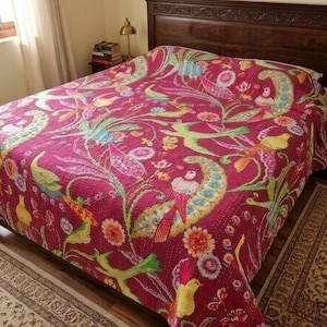 Op de afbeelding: Een magenta quilt met een levendig bloemen- en vogelpatroon. Het ontwerp bevat groene, gele, blauwe en roze vogels en bloemen. De quilt is uitgespreid over een houten bedframe, waardoor een kleurrijke en uitnodigende slaapkamer ontstaat.