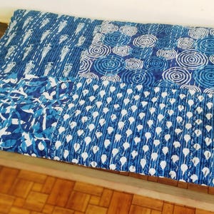 May include: A rectangular quilted throw blanket featuring a patchwork design of various blue and white block-printed patterns, including concentric circles, teardrops, and botanical motifs, finished with visible white running stitch quilting.