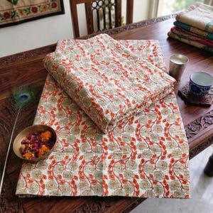 May include: Two folded floral patterned quilts in white, orange, and green sit on a dark wooden table. A small bowl of dried flowers and a peacock feather are also on the table. A cup and a candle are also visible.