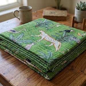 May include: A folded green quilt with a jungle design, featuring leopards, toucans, and tropical foliage. The quilt is stacked on a wooden table, showcasing its detailed pattern and vibrant colours.