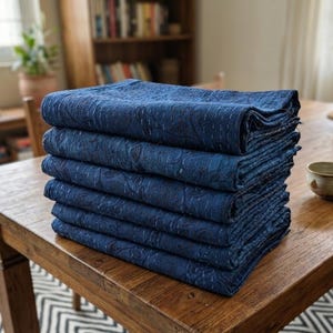 May include: A stack of six folded indigo blue quilts featuring a subtle tonal floral pattern and visible white running stitch embroidery throughout the fabric. The quilts are layered neatly on top of each other, showcasing the deep blue textile texture.