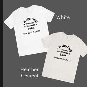 Peut inclure: Deux t-shirts avec le texte "I'm Writing Someone's Future Favorite Book How Cool Is That?" imprim&eacute; dessus. Un t-shirt est blanc et l'autre est en ciment chin&eacute;.