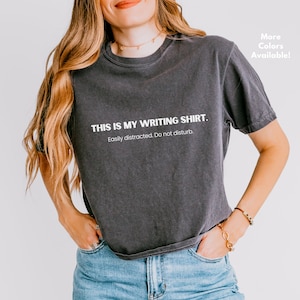 Peut inclure: Un t-shirt gris foncé avec le texte "THIS IS MY WRITING SHIRT. Easily distracted. Do not disturb." imprimé en blanc.