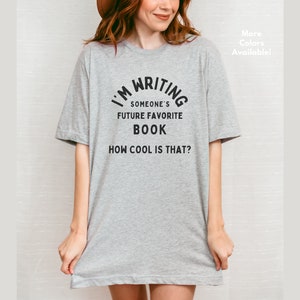 Peut inclure: T-shirt gris avec un texte noir qui dit "J'&eacute;cris le futur livre pr&eacute;f&eacute;r&eacute; de quelqu'un. C'est cool, non ?"