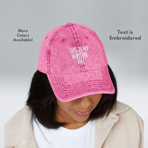 May include: A pink baseball cap with the embroidered text "THIS IS MY WRITING HAT." The hat is a faded pink colour and has a curved brim. The text "Text is Embroidered" is also visible.
