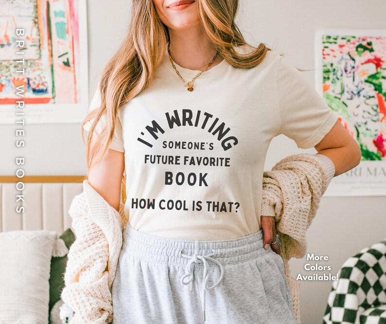 Peut inclure: Un t-shirt blanc avec du texte noir qui dit "J'&eacute;cris le futur livre pr&eacute;f&eacute;r&eacute; de quelqu'un. C'est cool, non ?"