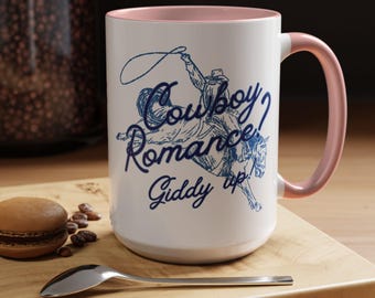 May include: White ceramic mug with a pink interior and handle. The mug features a blue illustration of a cowboy on a horse with the words "Cowboy Romance! Giddy up." A macaron and spoon are on a wooden surface.