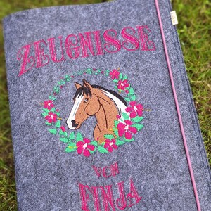 Könnte beinhalten: Grauer Filz-Dokumentenhalter mit rosa gesticktem Text und einem Pferdekopf-Design. Der Text lautet "ZEUGNISSE" über einem Pferdekopf, der von einem Blumenkranz umgeben ist, und "VON FINJA" darunter. Ein rosa Kordel verläuft am Rand.