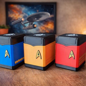 Könnte beinhalten: Drei quadratische Aufbewahrungsbehälter in Blau, Gelb und Rot, jeweils mit einem schwarzen Deckel und einem goldenen Delta-Emblem im Starfleet-Stil auf der Vorderseite. Die Behälter haben ein strukturiertes Muster am unteren Rand und ein kleines Emblem.