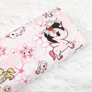 Op de afbeelding: Roze stof met een herhalend patroon van witte eenhoorns, roze kersenbloesems en roze sterren. De eenhoorns hebben zwarte manen en staarten en sommige hebben gouden accenten.