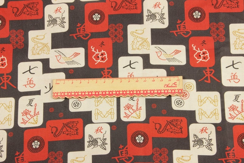 May include: Fabric with a repeating pattern of red and white tiles, each featuring Asian-inspired symbols and illustrations. The background is a dark gray. A ruler with red trim is placed across the fabric.