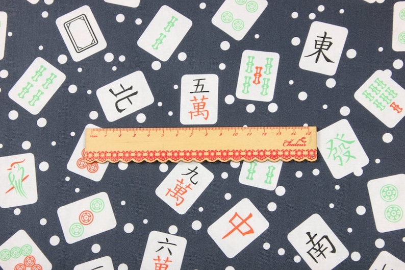 Pu&ograve; includere: Un tessuto blu scuro con un motivo a pois bianchi e un motivo ripetuto di tessere di mahjong bianche con caratteri cinesi e simboli di bamb&ugrave; verdi. Un righello &egrave; posizionato sopra il tessuto.