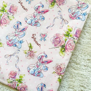 May include: Pale pink fabric printed with a repeating pattern of Sylveon characters, pink roses, bows, and hearts. The word "Sylveon" is written in cursive. The background has small white polka dots. The fabric is folded.