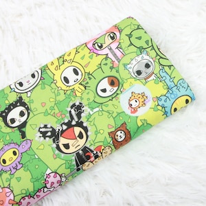May include: A green fabric with a repeating pattern of cartoon characters and cacti. The characters are colourful and include a black cat, a yellow bunny, a green monster, a red and black punk rock character, and a blue and yellow smiley face.