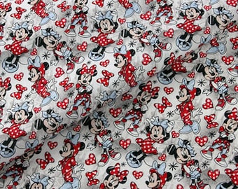Op de afbeelding: Witte stof met een herhalend patroon van rode en witte stippen Minnie Mouse-figuren. De achtergrond is lichtgrijs met witte stippen.