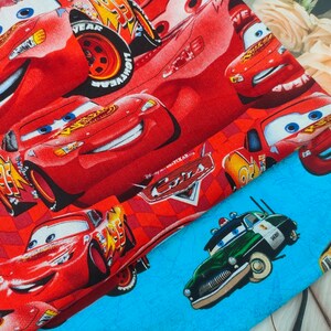 May include: Red fabric with a repeating pattern of cartoon racing cars, including Lightning McQueen. The fabric is folded over a blue fabric with a cartoon police car.