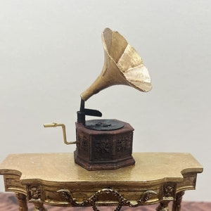 Könnte beinhalten: Ein goldfarbener Miniaturtisch mit einem verzierten Design und ein Miniatur-Grammophon mit einem goldfarbenen Horn auf der Oberseite. Tisch und Grammophon stehen auf einem braunen Holzboden.