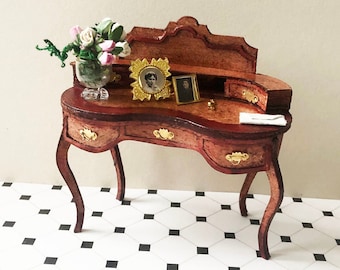 May include: A miniature wooden desk with a curved front, three drawers, and gold accents. The desk has a brown finish and is decorated with a vase of pink flowers, two framed photos, and a small gold object. The desk is sitting on a black and white checkered floor.
